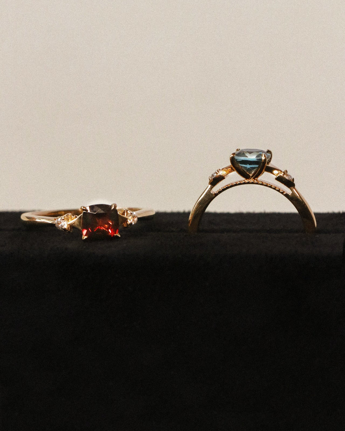 Two gold rings displayed on a black velvet surface against a light gray background. The left ring features a cushion garnet, and the right ring features a cushion topaz.