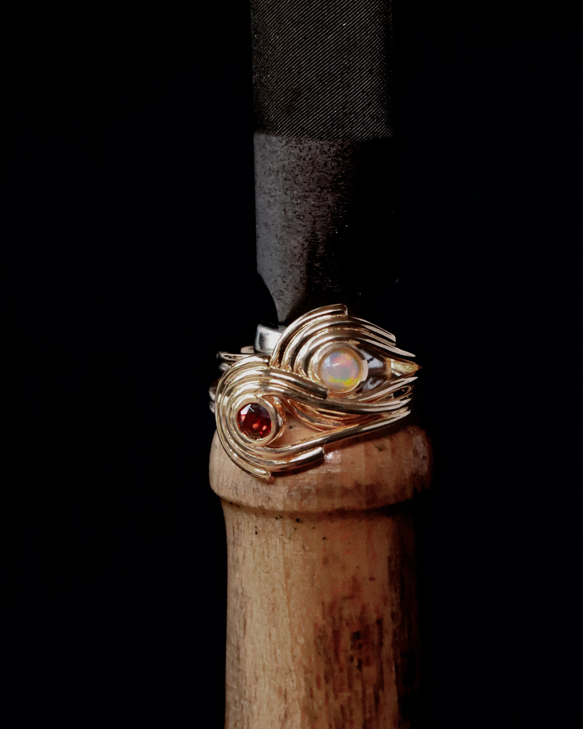 A close-up of a handmade wire-wrapped ring on a wooden handle against a solid black background. The ring features a gold-toned wrap with a red gemstone and a white iridescent opal-like stone.