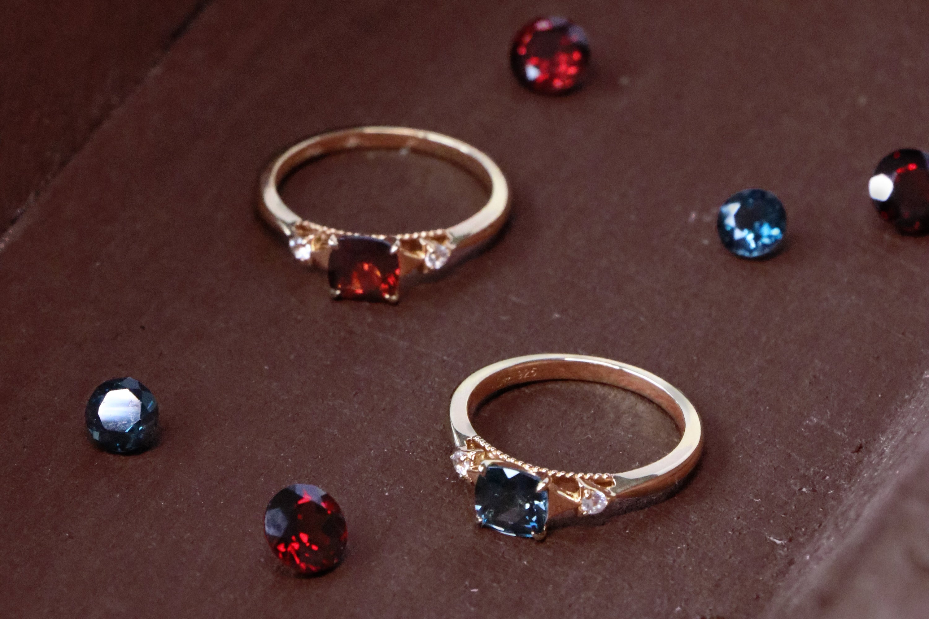 Two gold rings with marquise-cut gemstones, one featuring a red stone and the other a blue stone, displayed on a brown velvet jewelry box surrounded by loose matching gemstones.