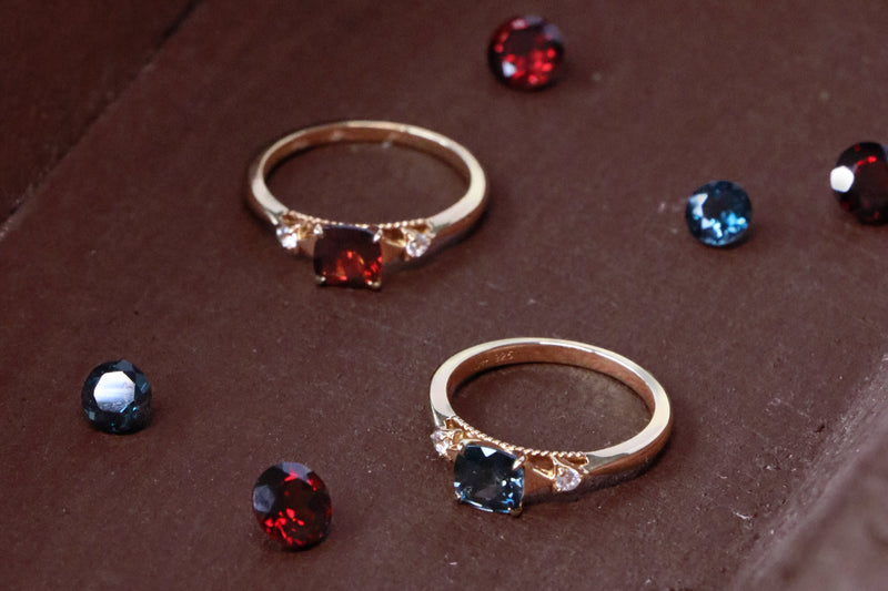 Two gold rings with marquise-cut gemstones, one featuring a red stone and the other a blue stone, displayed on a brown velvet jewelry box surrounded by loose matching gemstones.