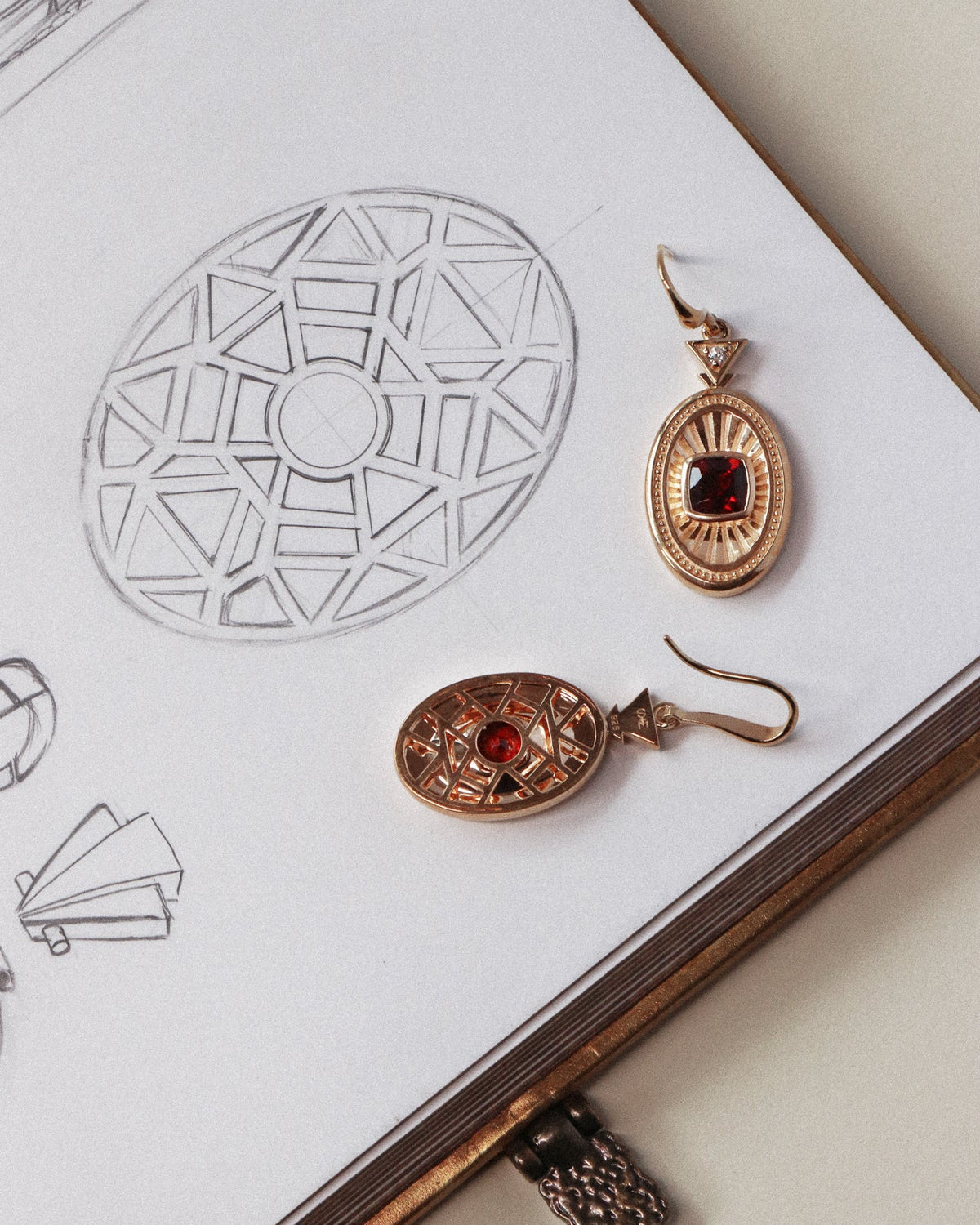 Two oval drop earrings with gold-toned filigree frames and garnet centers, displayed on an open sketchbook with geometric drawings.