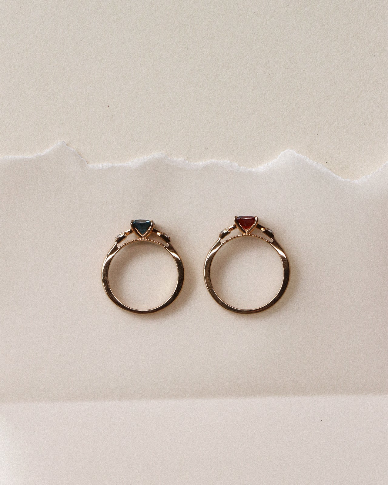 Two gold rings with small triangular gemstones, London blue topaz and red garnet, displayed side by side on a textured off-white surface.