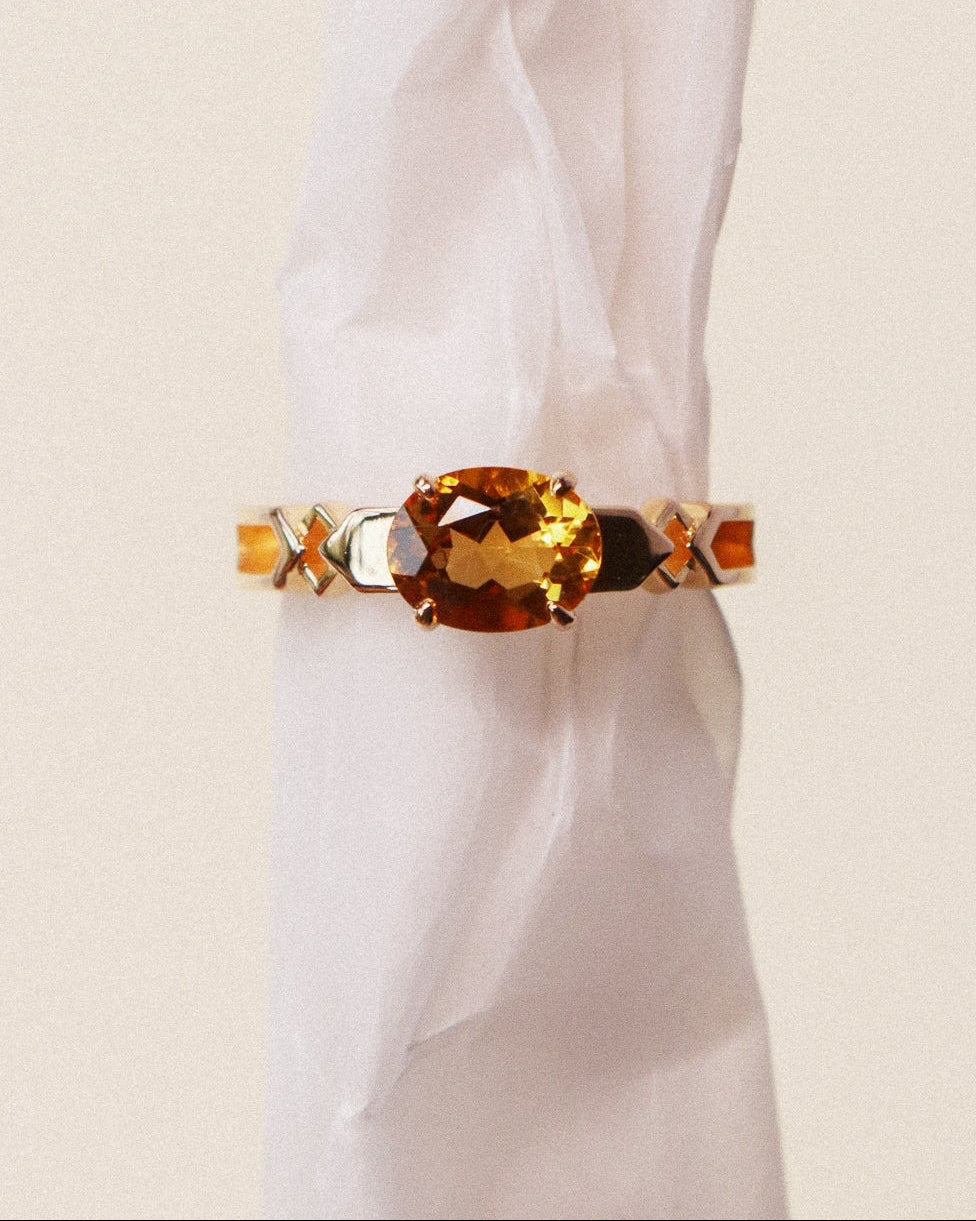A gold ring with a large oval amber citrine and small geometric side accents, displayed on a white fabric ring holder against a light beige background.